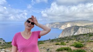 Yvonne Heckmann trägt ein pinkes T-Shirt, während im Hintergrund ein Ozean und Felsen zu sehen sind.