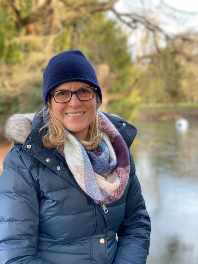 vonne Heckmann trägt eine blaue Jacke und eine blaue Wollmütze. Im Hintergrund sind Wasser und Bäume zu sehen.