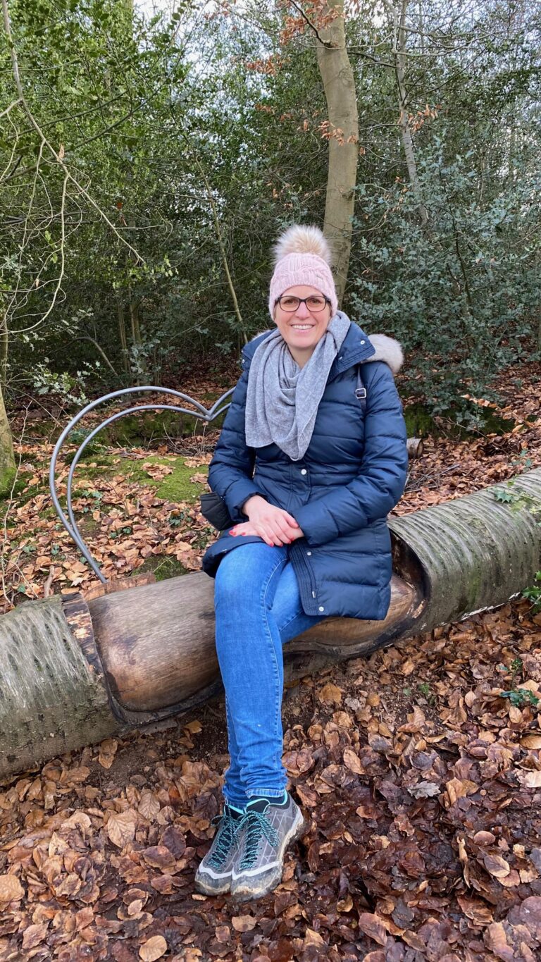 Yvonne Heckmann trägt eine blaue Winterjacke und rosa Bommelmütze und sitzt auf einem Baumstamm im Wald.