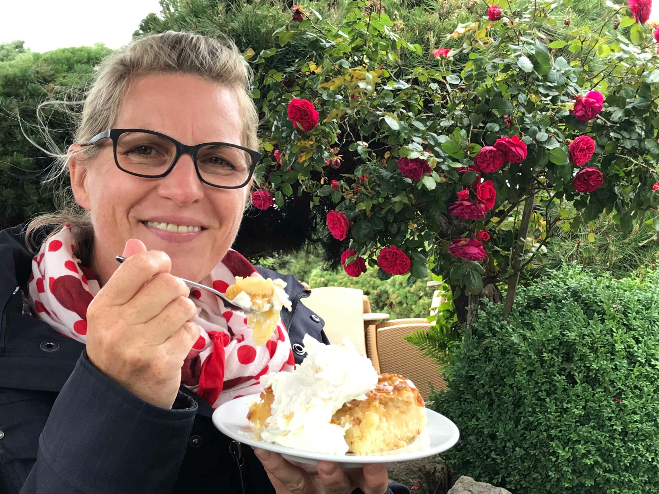 Yvonne Heckmann ist ein Bienenstich mit Sahne, während im Hintergrund ein Rosenbusch mit roten Rosen zu sehen ist.