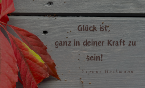 Ein herbstlich verfärbtes Ahornblatt auf grauem Holz mit dem inspirierenden Spruch 'Glück ist, ganz in deiner Kraft zu sein! - Yvonne Heckmann'.
