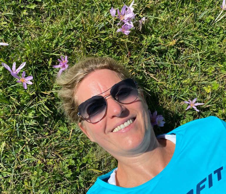 Yvonne Heckmann liegt glücklich, lachend auf einer grünen Wiese mit lila Blumen.