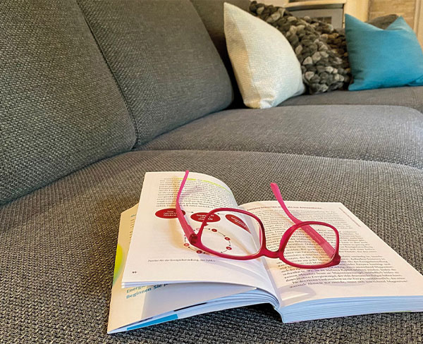 Ein graues Sofa mit einem aufgeschlagenen Buch, auf dem eine pinke Brille liegt."